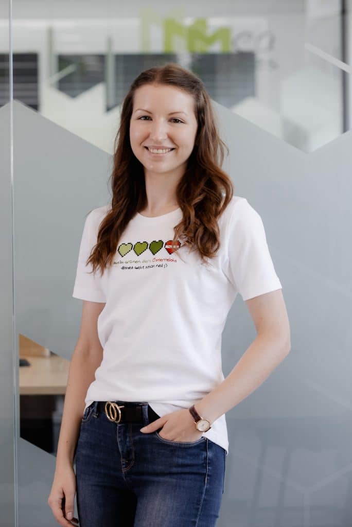 Kerstin Greifensteiner steht vor einer Glaswand und trägt ein weißes Shirt mit drei grünen Pixelherzen und einem roten Pixelherz.