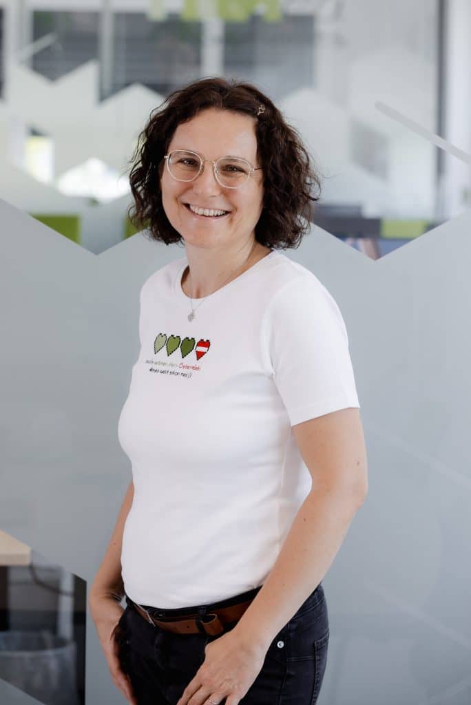 Maria Rücker-Ulm steht vor einer Glaswand und trägt ein weißes Shirt mit drei grünen Pixelherzen und einem roten Pixelherz.