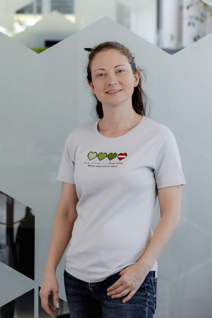 Beatrix Töglhofer steht vor einer Glaswand und trägt ein weißes Shirt mit drei grünen Pixelherzen und einem roten Pixelherz.