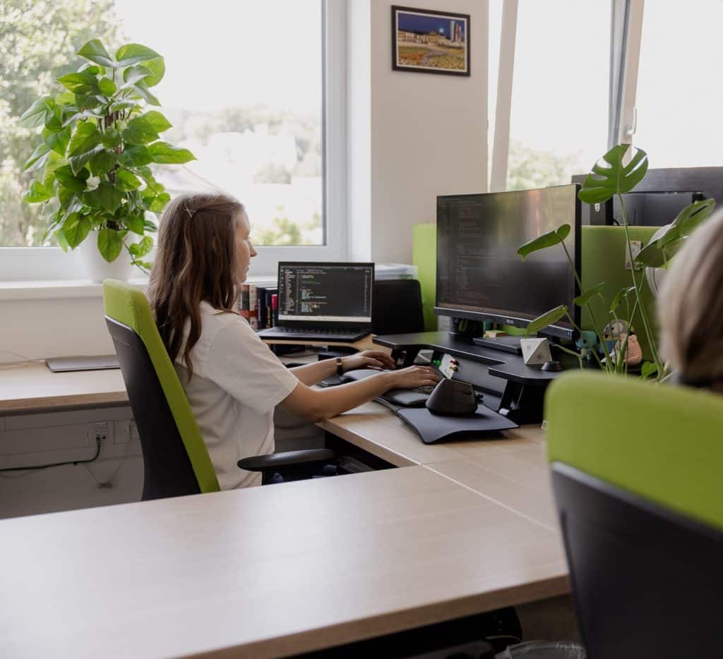 Man sieht eine Frau von schräg hinten, wie sie an ihrem Arbeitsplatz Code schreibt. Vor ihr befinden sich ein großer Monitor, ein Laptop und eine Tastatur