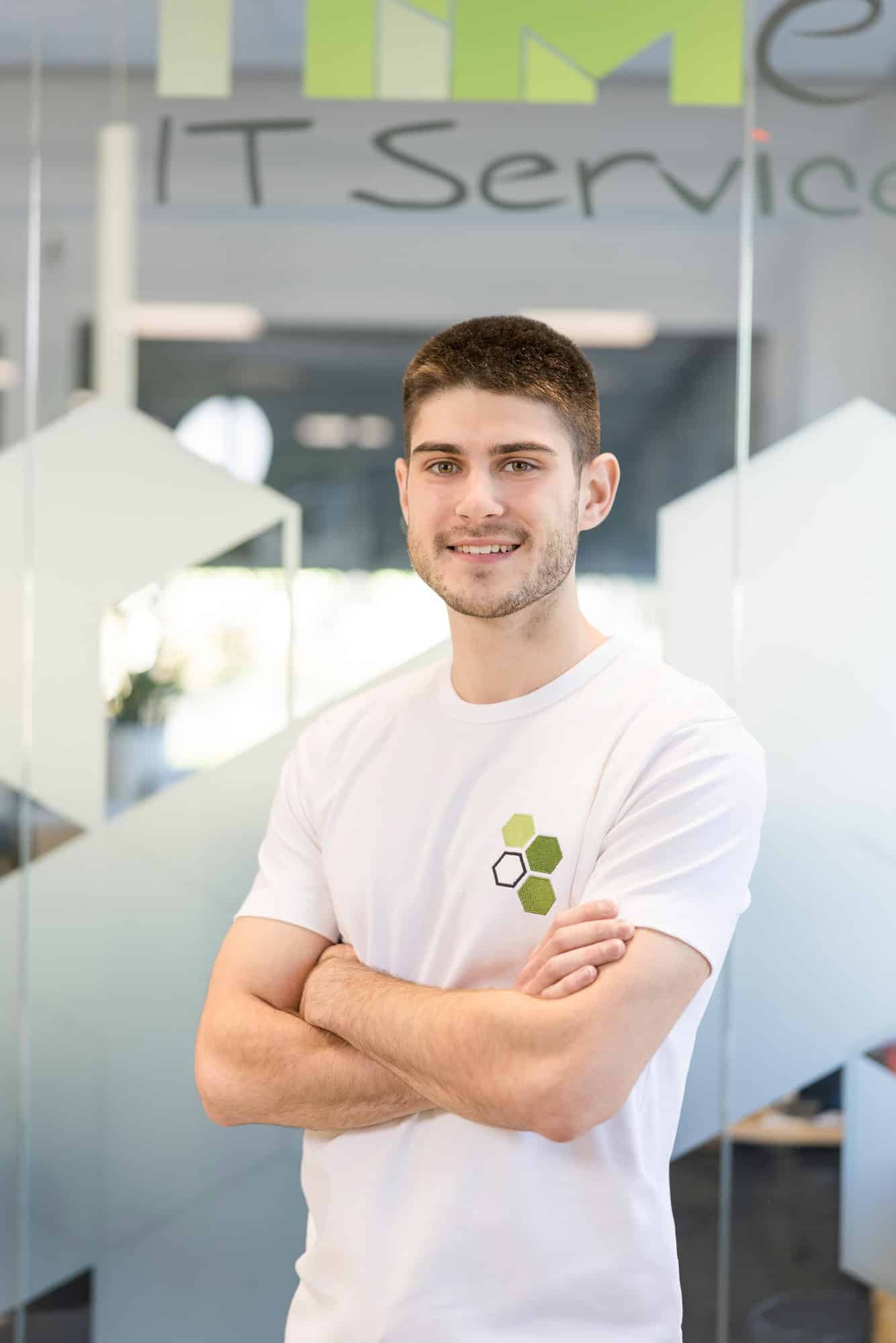 Lukas Url Lukas Url steht vor einer Glaswand und trägt ein weißes Shirt mit einer Applikation aus drei grünen Waben des mea IT Logos und einer schwarz umrahmten Wabe.
