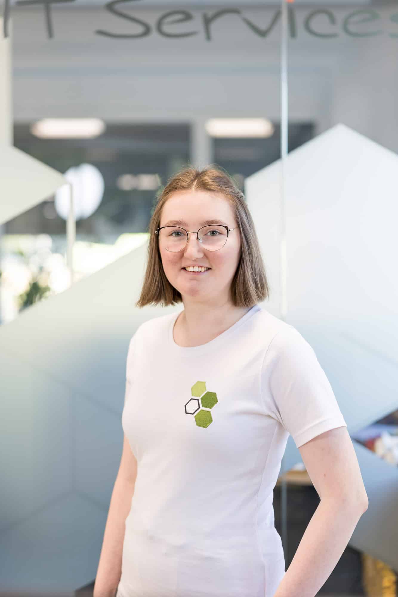Laura Priesch Laura Priesch steht vor einer Glaswand und trägt ein weißes Shirt mit einer Applikation aus drei grünen Waben des mea IT Logos und einer schwarz umrahmten Wabe.